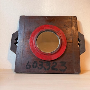 May include: A rustic, square wooden mirror with a circular mirror set in a red painted frame. The wooden frame is dark brown with a distressed finish and has decorative side handles. Black numbers "603323" are painted on the wood.