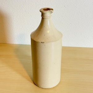 May include: A tall, cylindrical stoneware bottle in a light beige color. The bottle features a narrow neck with a distinct, slightly flared rim and a small pouring spout. The surface has a smooth, matte finish with subtle speckling throughout.