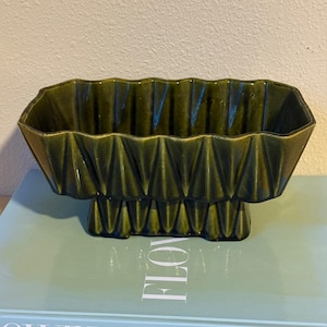 May include: A dark green ceramic planter with a geometric design. The planter has a rectangular shape with a series of triangular facets around the rim and base. The planter is elevated on a pedestal base.