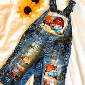 May include: Blue denim overalls with distressed details and patches featuring farm scenes. The overalls have adjustable straps and a classic overall design. Two yellow sunflowers are placed near the overalls.