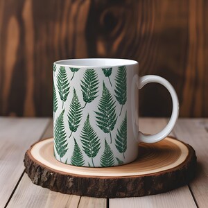 May include: White ceramic mug with a green fern leaf pattern. The mug is sitting on a wooden coaster.