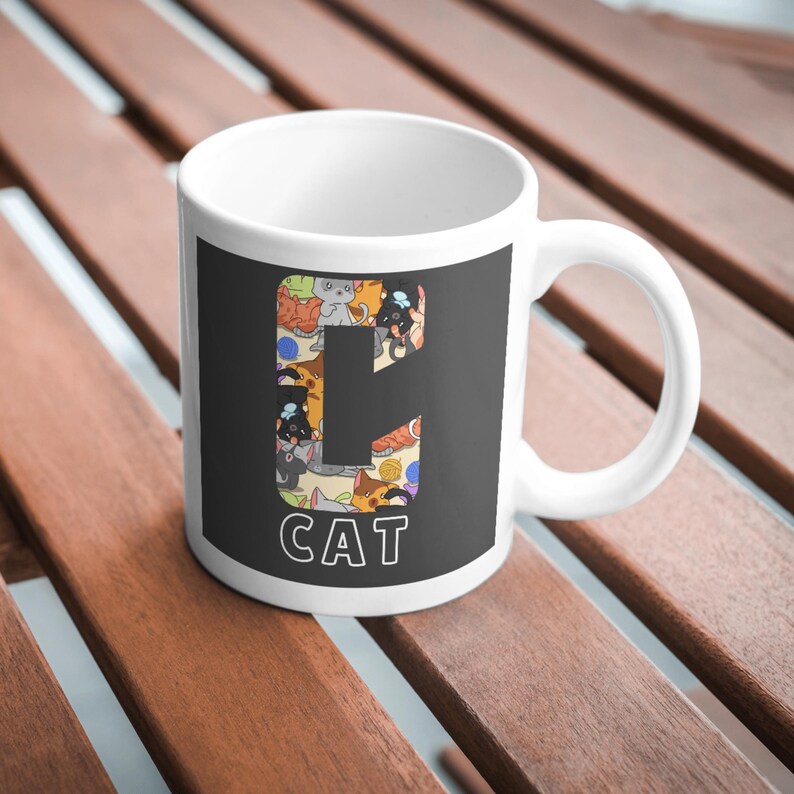 May include: White ceramic mug with a black panel featuring the letter "C" filled with cat illustrations in various colors. The word "CAT" is printed in white below the letter. The mug has a curved handle.