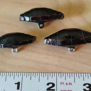 May include: Three black, vintage-style buttons with a textured, segmented design. Each button has a silver-colored metal loop on the underside. The buttons are arranged on a wooden surface next to a ruler.