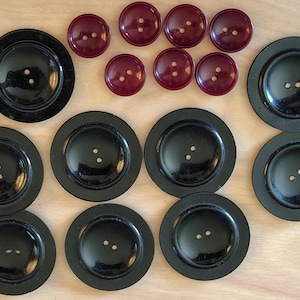 May include: An assortment of black and burgundy buttons. The black buttons are large and round with two holes, while the burgundy buttons are smaller and also have two holes. The buttons are arranged on a light wooden surface.