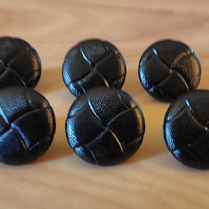 May include: A set of six round black buttons featuring a textured surface with a cross-hatched pattern. Each button has a domed shape and a smooth, glossy finish, arranged in two rows of three on a light-colored surface for clear visibility.