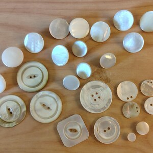 May include: A collection of various sizes and shapes of iridescent, off-white buttons. The buttons include round, oval, and square shapes, with two or four holes for sewing. Some buttons have a pearlescent sheen.