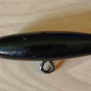 May include: A black, oblong wooden toggle with a metal ring attached. The toggle is smooth and glossy, with visible wear and tear. The metal ring is tarnished, showing signs of age and use. The item is set against a light brown wooden surface.