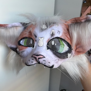 May include: A handmade cat mask with a white, orange, and black color scheme. The mask features large green eyes, a pink nose, and white whiskers. The ears are a light orange color with white fur.