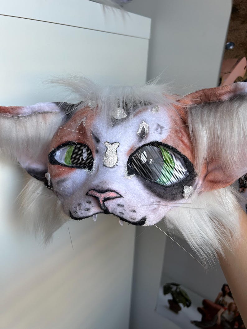 May include: A handmade cat mask with a white, orange, and black color scheme. The mask features large green eyes, a pink nose, and white whiskers. The ears are a light orange color with white fur.