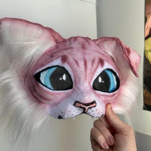 May include: A pink and white cat mask with large blue eyes and mesh eye coverings. The mask has a detailed, textured appearance with white fur around the face and ears. The mask is held by a hand.