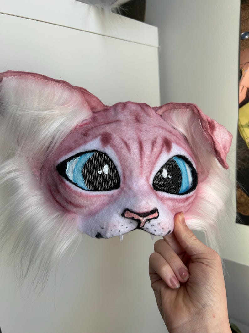 May include: A pink and white cat mask with large blue eyes and mesh eye coverings. The mask has a detailed, textured appearance with white fur around the face and ears. The mask is held by a hand.