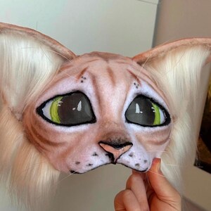 May include: A cat mask with large, expressive eyes and a detailed, realistic design. The mask features a light brown and tan color scheme with green eyes and white fur around the ears. The mask is held by a hand.
