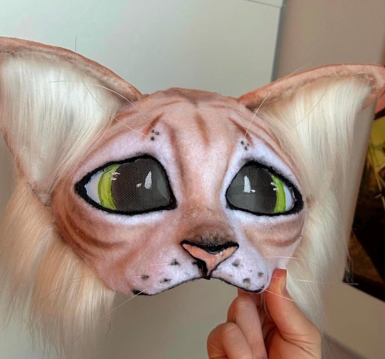 May include: A cat mask with large, expressive eyes and a detailed, realistic design. The mask features a light brown and tan color scheme with green eyes and white fur around the ears. The mask is held by a hand.