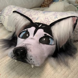 May include: A plush husky dog head with black and white fur, large blue eyes, and a black nose. The ears are black with white fur inside. The plush has small white fangs and is lying on a patterned surface.