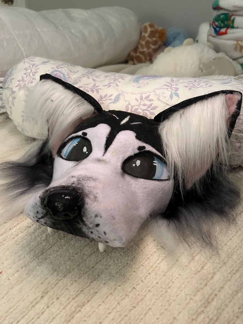 May include: A plush husky dog head with black and white fur, large blue eyes, and a black nose. The ears are black with white fur inside. The plush has small white fangs and is lying on a patterned surface.