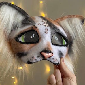 May include: A handmade cat mask with a realistic design. The mask features a white, brown, and black color scheme, with green eyes and a pink nose. The mask has long, fluffy, white and tan fur around the ears and face.