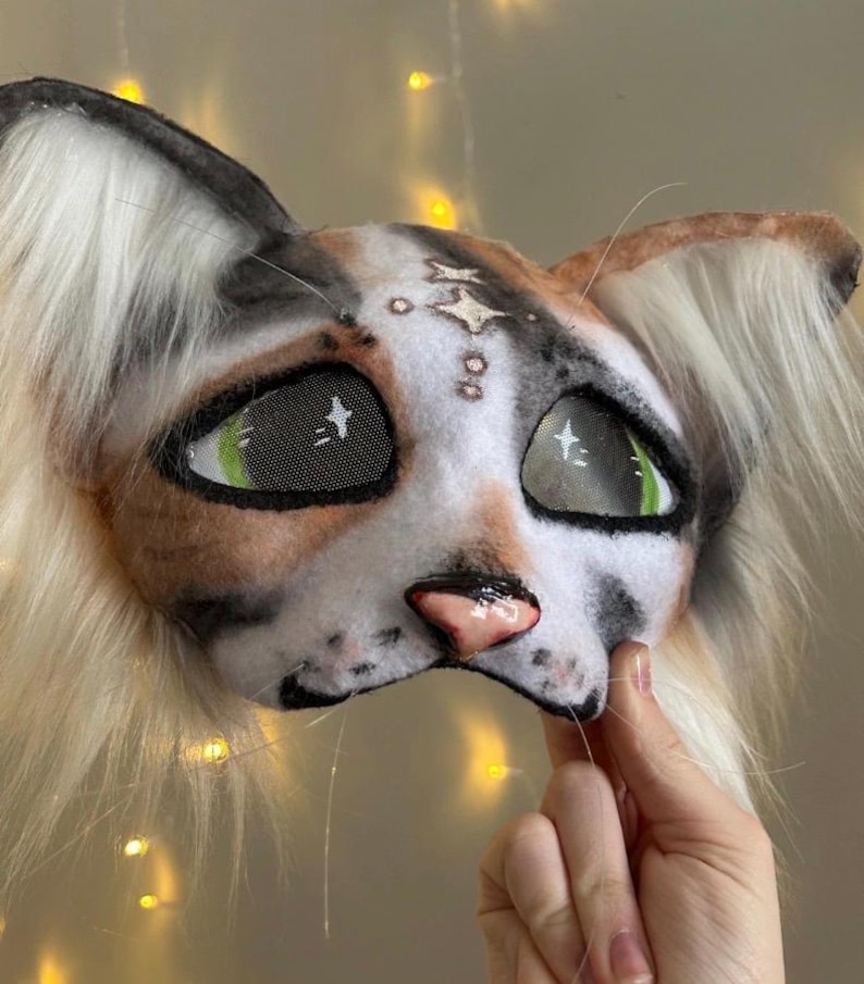 May include: A handmade cat mask with a realistic design. The mask features a white, brown, and black color scheme, with green eyes and a pink nose. The mask has long, fluffy, white and tan fur around the ears and face.
