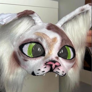 May include: A white and brown cat mask with large, green mesh eyes. The mask has long, white fur around the face and ears, a pink nose, and black detailing around the mouth and eyes. The mask is designed to be worn.