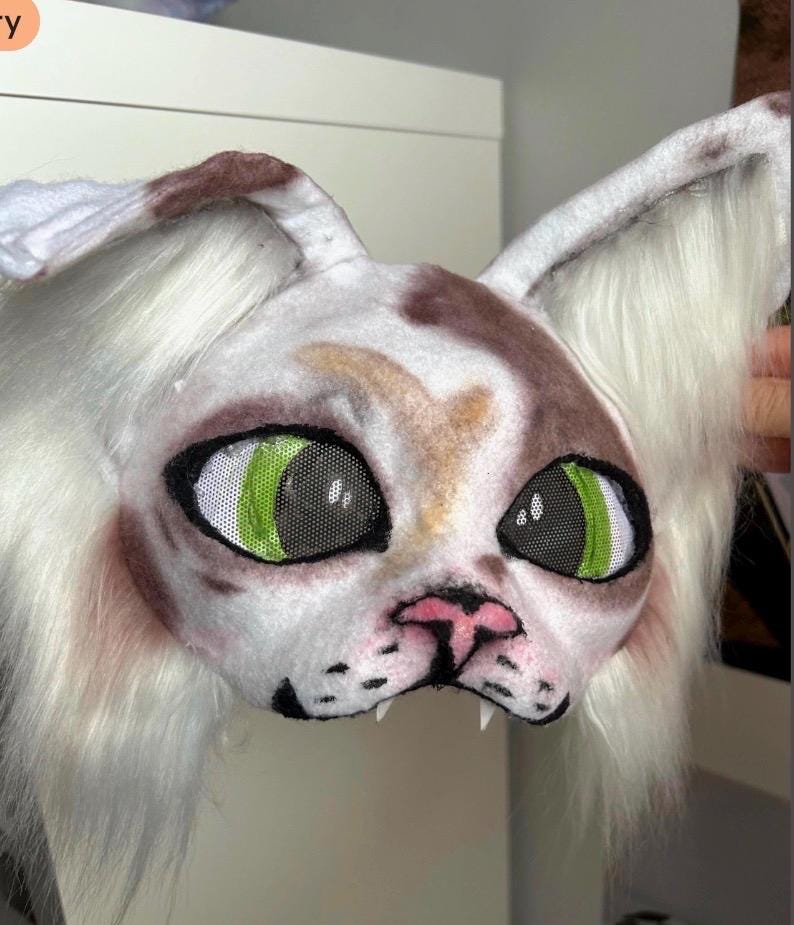 May include: A white and brown cat mask with large, green mesh eyes. The mask has long, white fur around the face and ears, a pink nose, and black detailing around the mouth and eyes. The mask is designed to be worn.