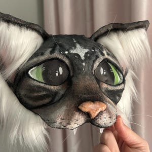 May include: A detailed cat mask with a black and gray face, green eyes, and a pink nose. The mask has white fur around the ears and cheeks, and visible whiskers. The mask is designed to be worn.