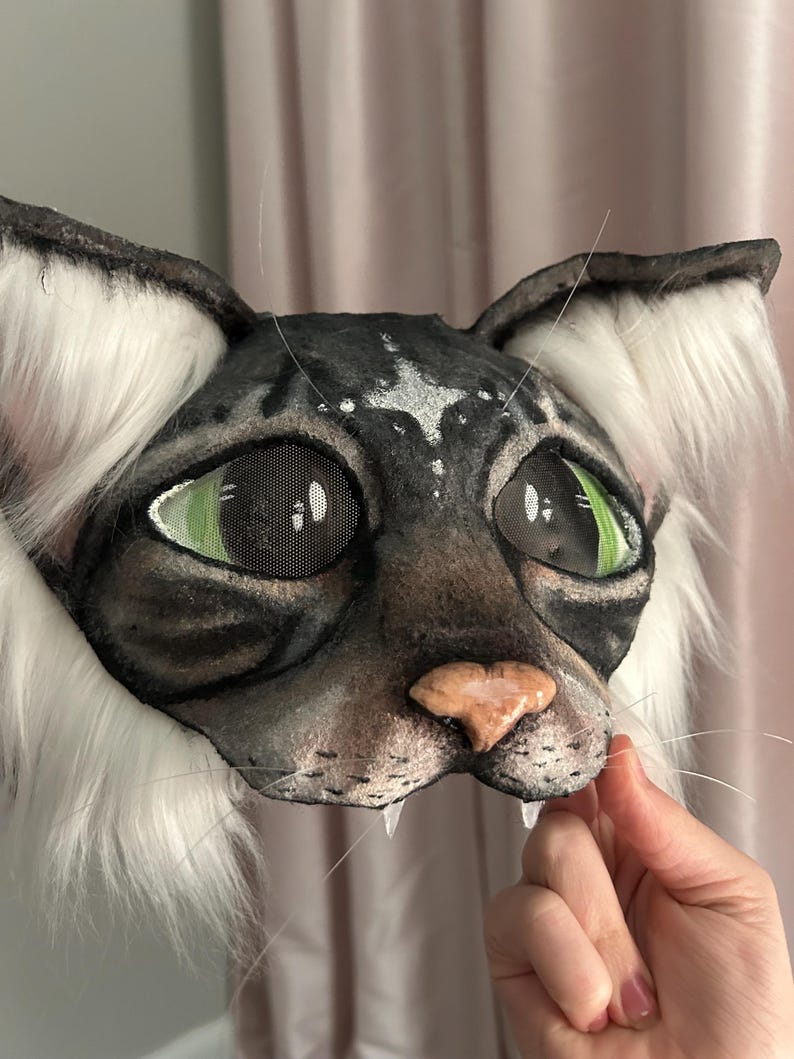May include: A detailed cat mask with a black and gray face, green eyes, and a pink nose. The mask has white fur around the ears and cheeks, and visible whiskers. The mask is designed to be worn.
