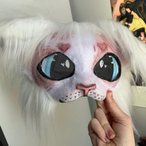 May include: A white and pink cat mask with large blue eyes and black mesh eye coverings. The mask has long white fur around the edges and is held by a hand.