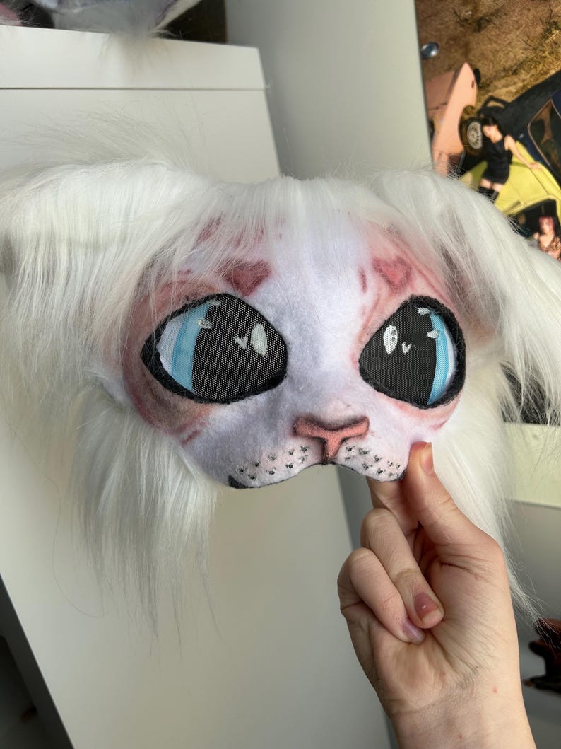 May include: A white and pink cat mask with large blue eyes and black mesh eye coverings. The mask has long white fur around the edges and is held by a hand.