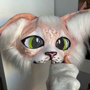 May include: A handmade cat mask with large, expressive eyes. The mask features a peach-colored face with white fur around the ears and cheeks. The eyes are black with bright green irises. The mask is held by a person wearing a gray sweatshirt.