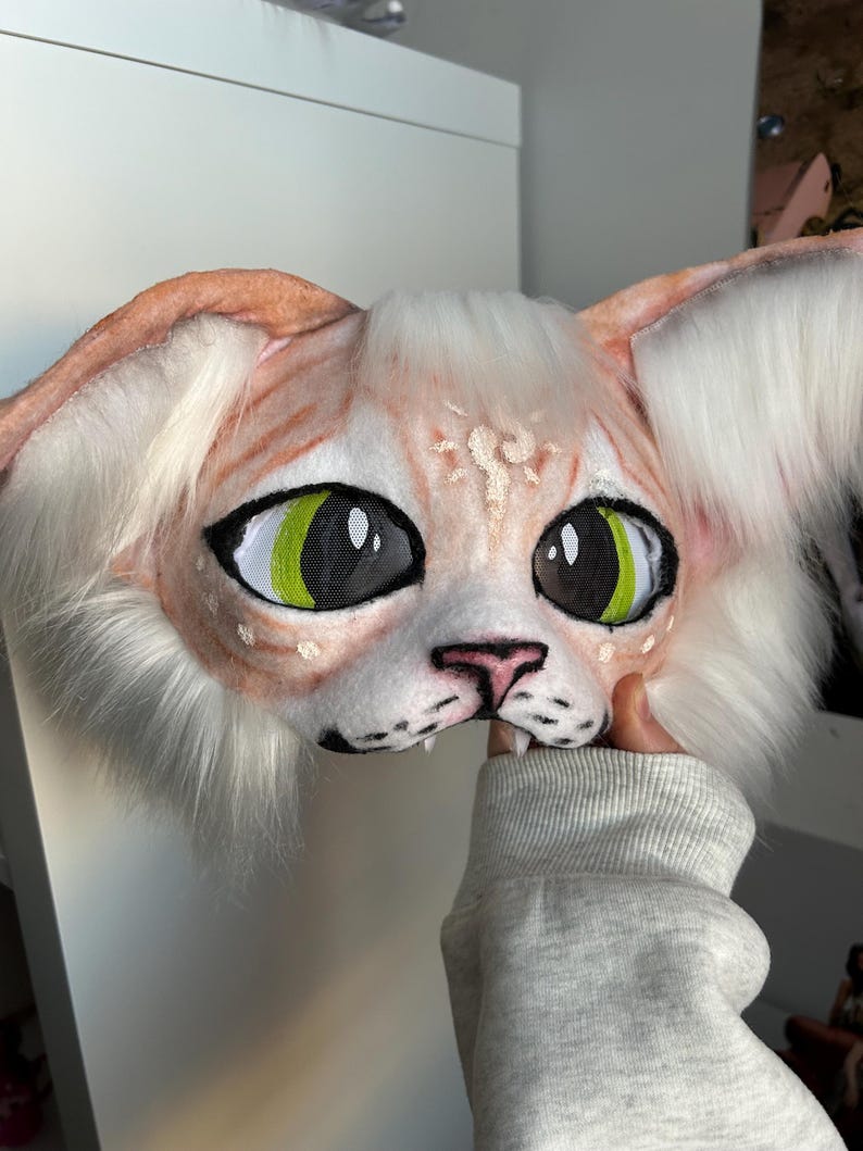 May include: A handmade cat mask with large, expressive eyes. The mask features a peach-colored face with white fur around the ears and cheeks. The eyes are black with bright green irises. The mask is held by a person wearing a gray sweatshirt.