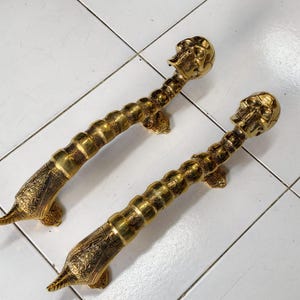 May include: A pair of gold-coloured metal door handles featuring a skull at the top end and a tapered, textured base. The handles have a segmented, cylindrical body with intricate engraved patterns along the surface and mounting brackets.