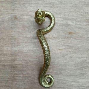 Peut inclure: Une poignée de meuble en forme de serpent, de couleur laiton. La poignée présente une tête de serpent détaillée avec une gueule ouverte et des crocs. Le corps du serpent est incurvé vers le bas avec un motif texturé, se terminant par une queue enroulée.