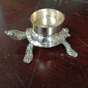 Könnte beinhalten: Ein silberner Kerzenhalter in Form einer Schildkröte mit einer runden, silbernen Oberseite. Der Halter ist aus Metall und hat ein detailliertes Design.