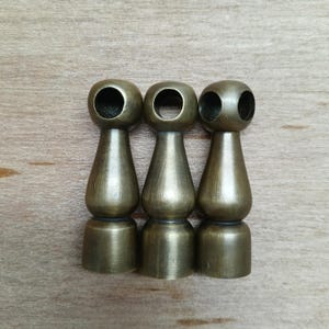 Puede incluir: Tres remates de metal de color dorado antiguo con una parte superior redonda y una base decorativa. Cada remate tiene una abertura circular en la parte superior.