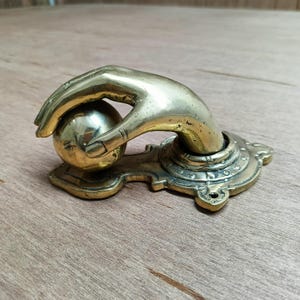 May include: A brass door knocker shaped like a hand holding a sphere. The knocker features an ornate design with intricate details.
