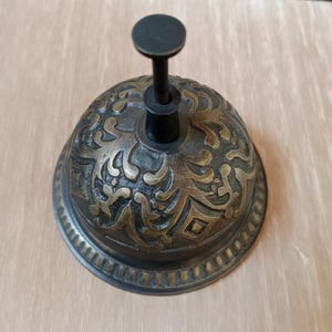 May include: Antique brass bell with ornate floral design. The bell has a dark handle and a round top. The bell is made of metal and has a vintage look. The bell is a decorative item.