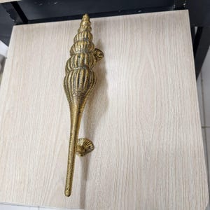 以下が含まれることがあります： 真鍮製の長い垂直ドアハンドルで、質感のあるメタリックな仕上げが施されています。ハンドルは螺旋状の貝殻の形をしており、先細りの尖った端と、取り付けブラケットが付いた幅の広いリブ付きの上部セクションが特徴です。