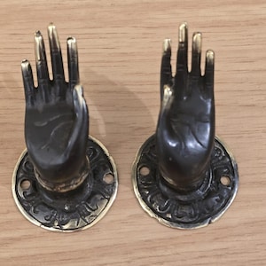 Könnte beinhalten: Zwei türgriffartige Handformen in Bronzefarbe mit goldfarbenen Fingerspitzen. Jeder Griff ist auf einer kreisförmigen Basis mit einem verzierten Design und einem goldfarbenen Rand montiert. Die Griffe sind auf einer hellbraunen Holzoberfläche angeordnet.