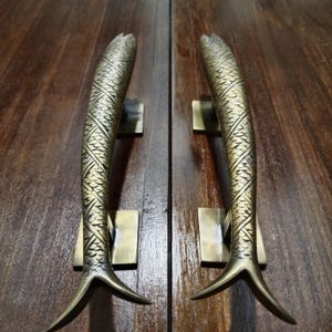 May include: A pair of antique brass door handles shaped like fish with intricate scale designs. The handles are mounted on a wooden door.
