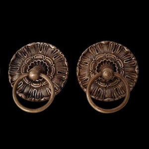 Puede incluir: Dos tiradores de armario antiguos de bronce con diseño floral. Cada tirador presenta una placa trasera circular con tallas detalladas en forma de pétalos y un asa redonda. Los tiradores están sobre un fondo negro.
