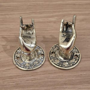 Könnte beinhalten: Zwei messingfarbene Türgriffe in Handform. Jeder Griff hat die Form einer Hand, bei der der Zeige- und kleine Finger ausgestreckt sind. Die Griffe sind auf runden Sockeln mit dekorativen Mustern montiert. Die Hardware ist für die Montage an einer Tür vorgesehen.