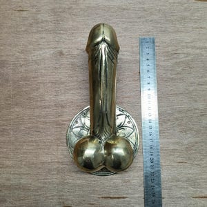 Puede incluir: Un llamador de puerta de latón con forma de figura masculina estilizada con un diseño floral. El llamador está unido a una base redonda con un diseño floral.