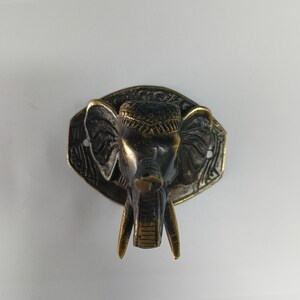 May include: A brass elephant head wall hook with intricate details. The elephant has a dark brown finish with gold accents. The hook is attached to a decorative plaque with a floral design.