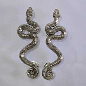 Peut inclure: Deux poignées de porte argentées en forme de serpents. Les poignées sont ornées d'écailles et ont une queue enroulée. Les têtes de serpent sont en haut, et les poignées sont montées sur des bases rondes argentées.