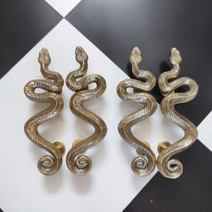 May include: Four brass-coloured snake-shaped door handles. Each handle has a detailed serpent design with a textured body and a coiled tail. The handles are mounted on a black and white checkered surface.