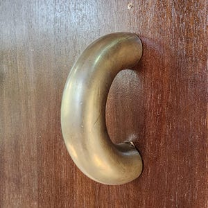 May include: A close-up of a brass door handle with a rounded, curved shape. The handle is mounted on a wooden door.