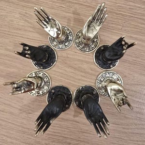Puede incluir: Una colección de manijas de puerta decorativas con forma de manos. Algunas son doradas, otras negras. Las manos están en varios gestos, incluyendo palmas abiertas y el signo de la mano 'rock on'. Cada manija está montada sobre una base circular decorativa.