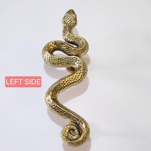Peut inclure: Une sculpture de serpent dorée avec un motif texturé en forme d'écailles. Le serpent est enroulé dans une pose sinueuse verticale, la tête levée. La sculpture a une finition polie et est posée sur un fond blanc.