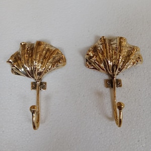 Può includere: Due ganci in ottone color oro a forma di conchiglie. I ganci sono montati su una parete bianca.