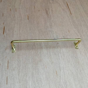 May include: A gold-toned metal towel bar with rounded ends and square brackets on each side. The bar is mounted on a light brown wooden surface.