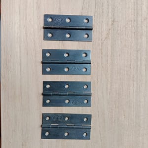 Könnte beinhalten: Vier schwarze Metallscharniere in verschiedenen Größen. Die Scharniere sind abgerundet und für Türen oder Schränke konzipiert. Die Scharniere sind vertikal auf einer Holzoberfläche angeordnet.